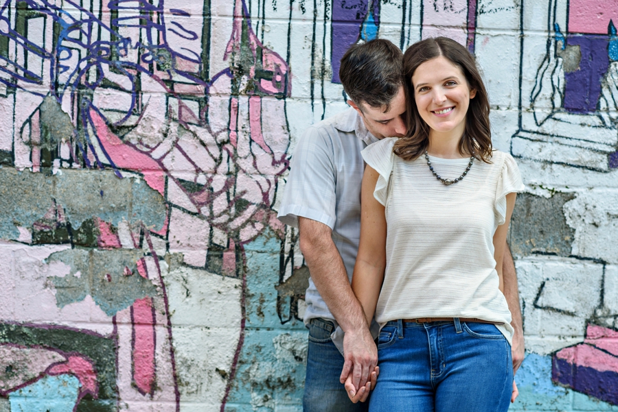 A cute couple stand in front of a graffiti wall of ben franklin in fishtown.